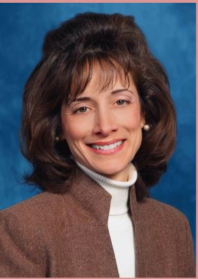 Woman with short brown hair and bangs, white skin, and wearing a brown top smiling.