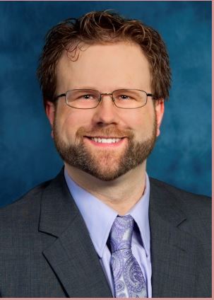 Man with curly brown hair, white skin, glasses, and a beard smiling with a blue suit.